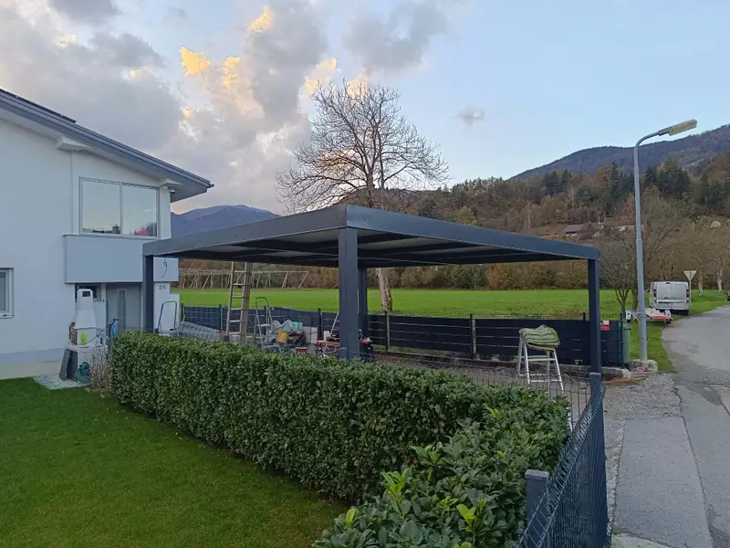 Samostoječ kovinski nadstrešek ob hiši, v ozadju travnik in hribi